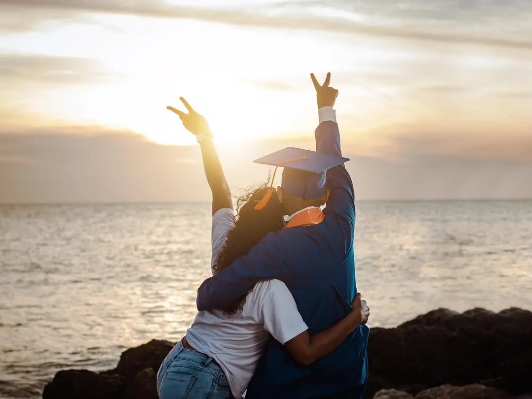 A graduate in cap and gown hugging a friend by the sea at sunset, both raising a hand